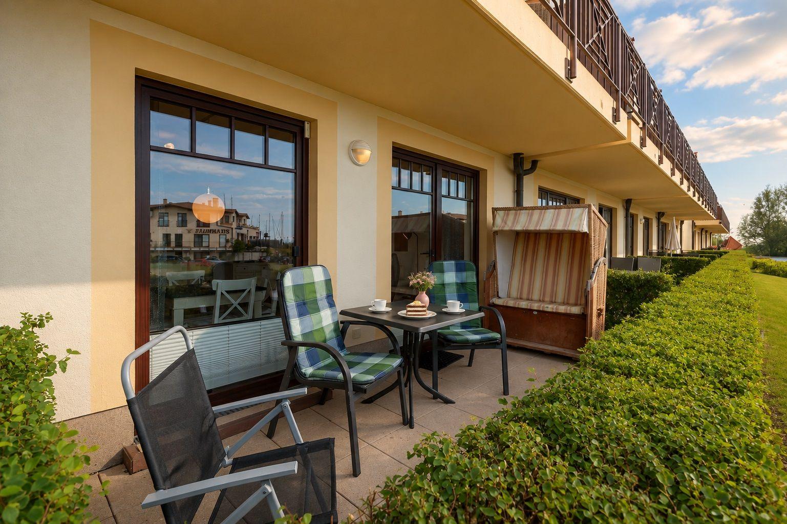 Terrasse mit Tisch, zwei Stühlen und Liegestuhl neben Haus mit Balkon.