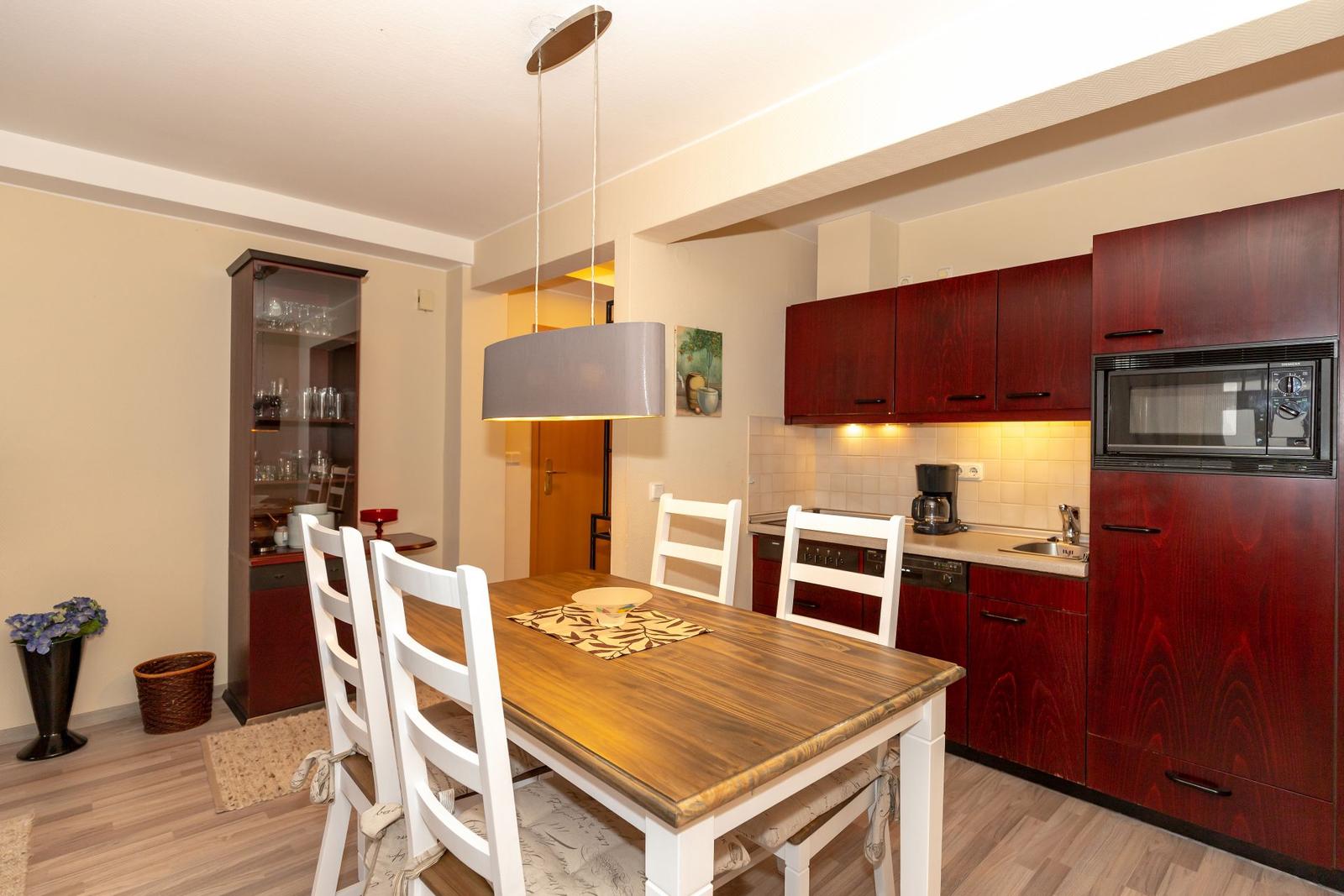 Dining area with wooden table and white chairs. Kitchen with dark cabinets and microwave.