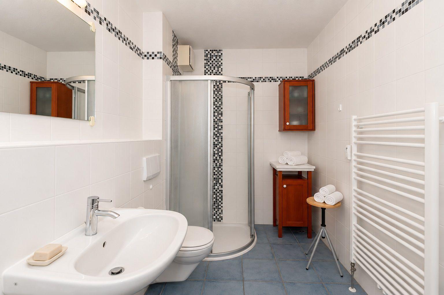 Badezimmer mit Dusche, Waschbecken und Toilette. Holzschränke und Heizkörper sind sichtbar.