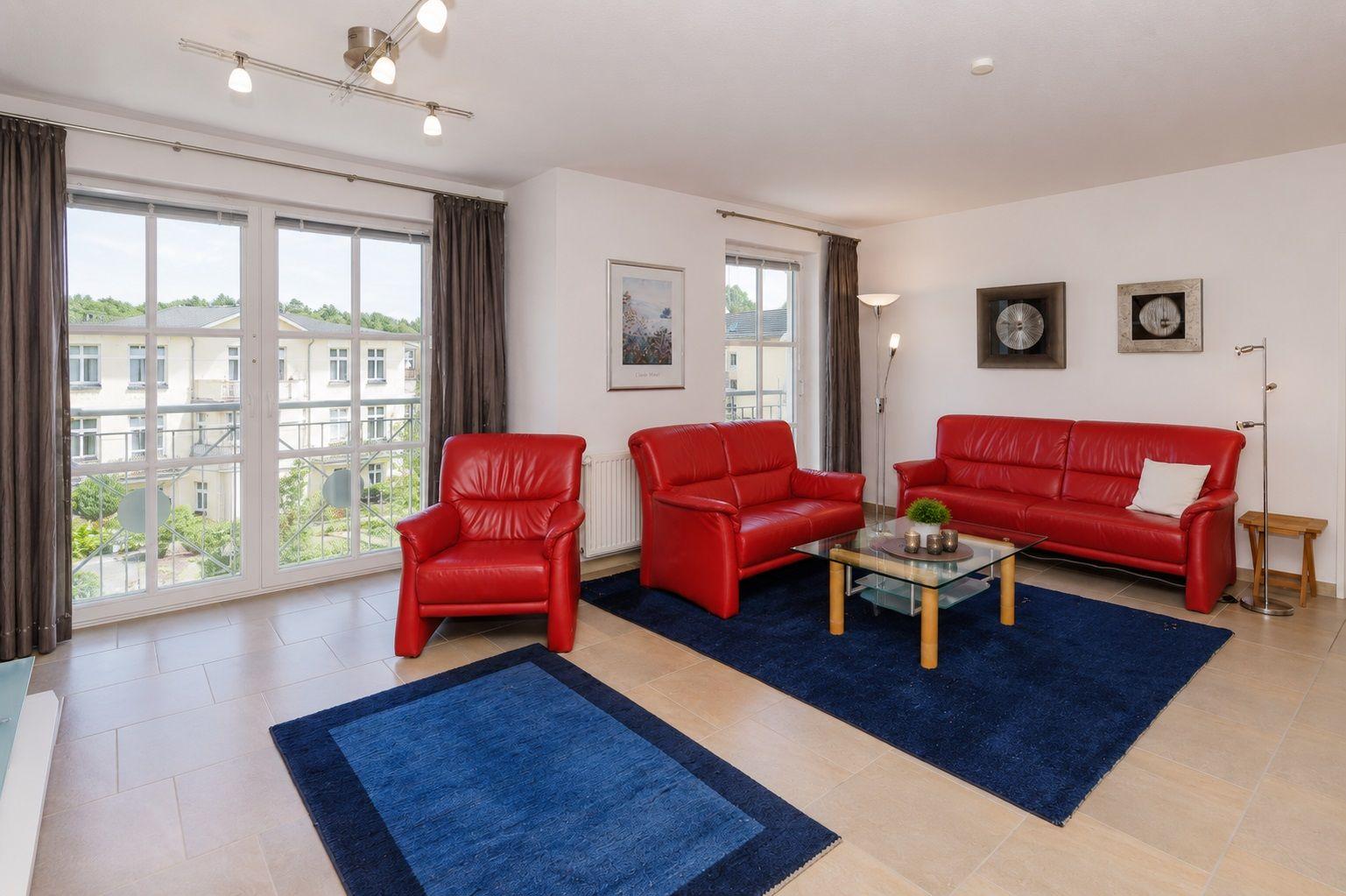 Wohnzimmer mit rotem Sofa, Sessel und blauem Teppich. Große Fenster und helle Wände.