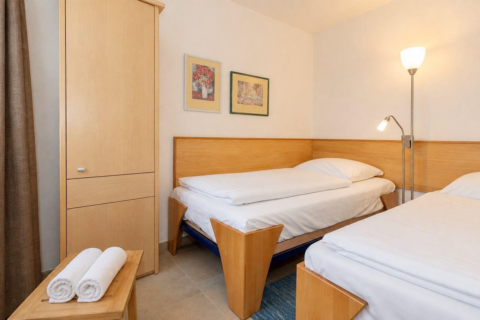 Zwei Betten mit weißer Bettwäsche, Holzgestell und Wandbilder. Stehlicht und Kleiderschrank.