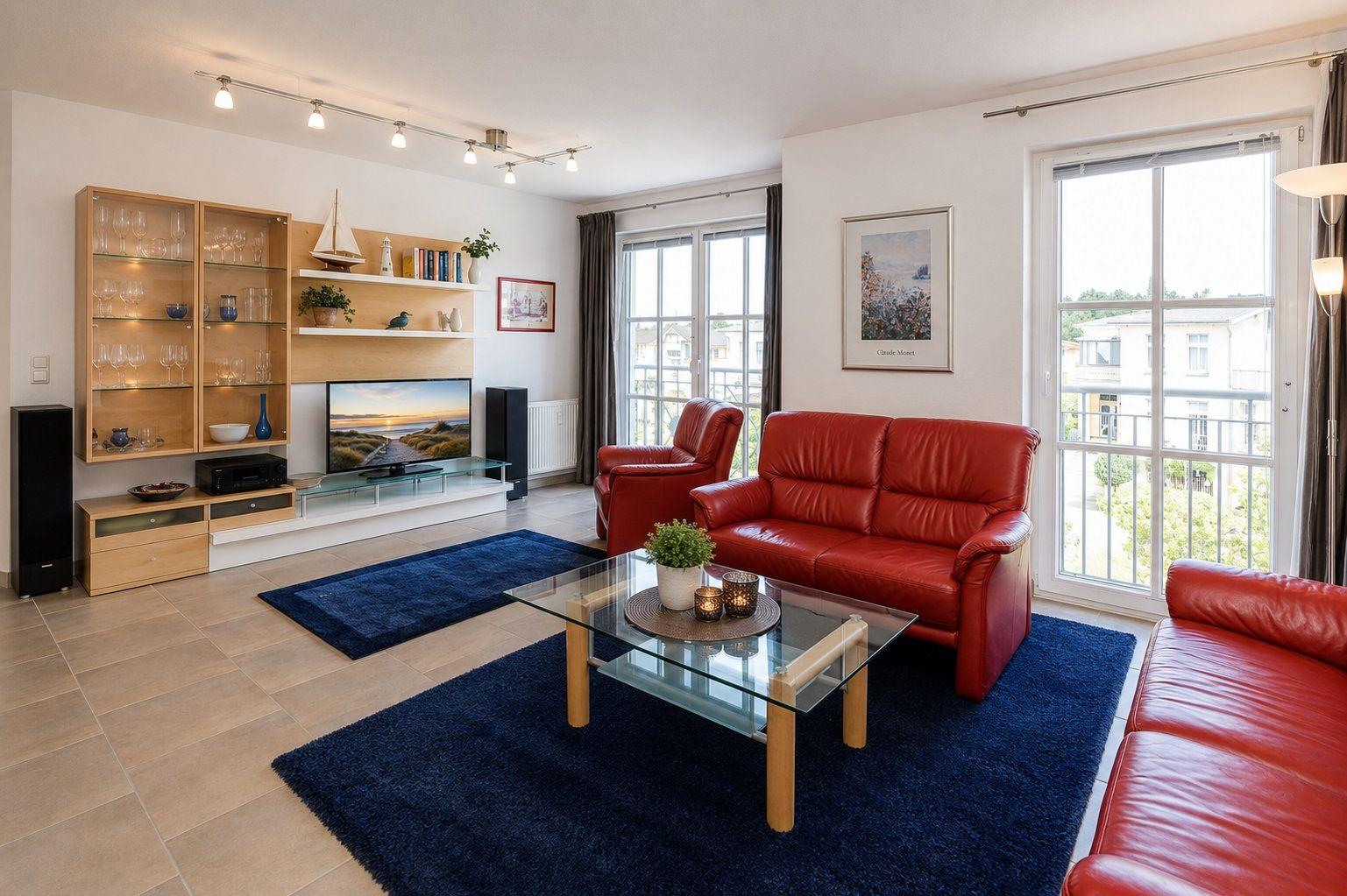 Wohnzimmer mit rotem Sofa, Glas-Tisch und blauem Teppich. Fenster mit Blick auf Balkon.