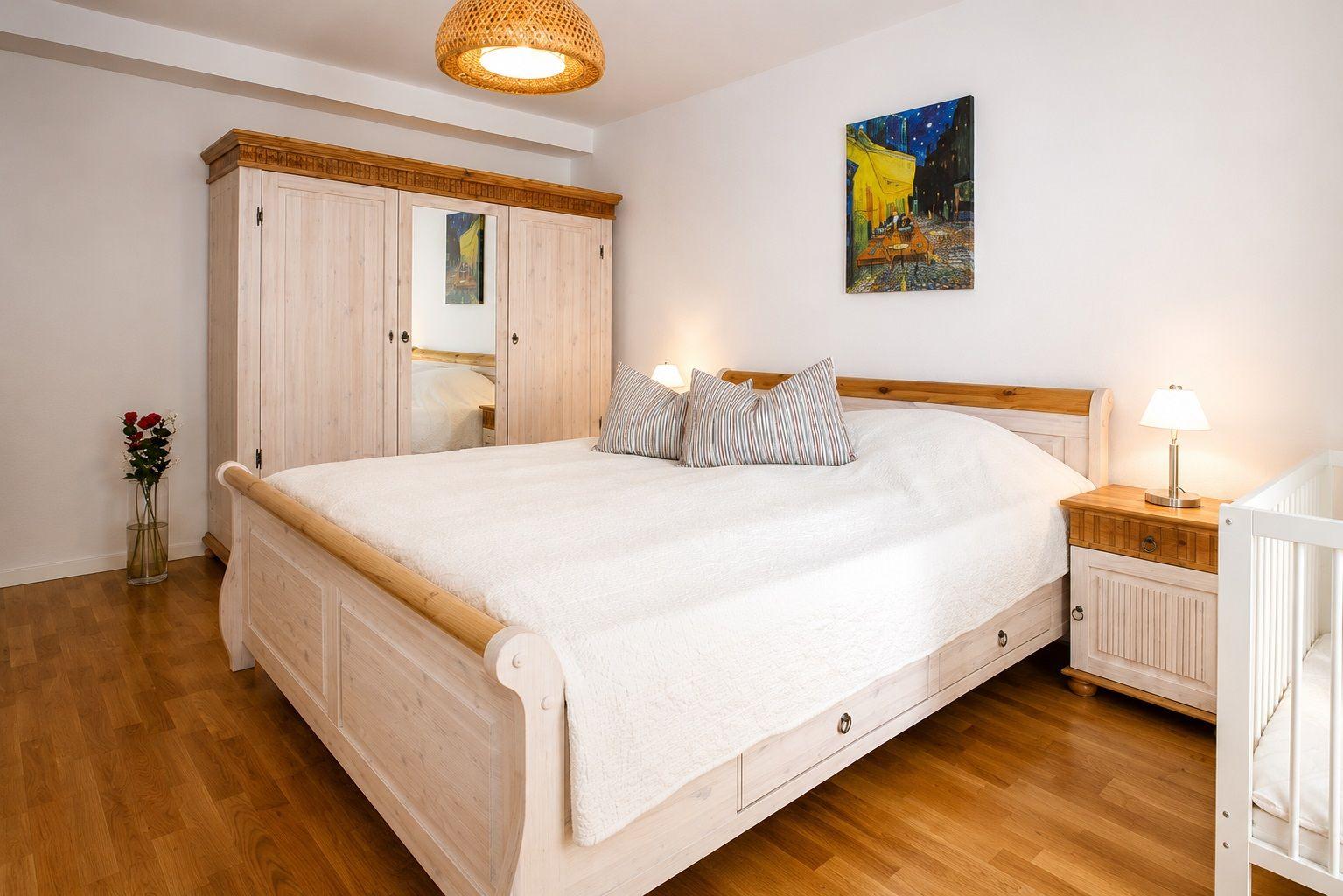 Schlafzimmer mit Bett, Kleiderschrank, Nachttisch und Kinderbett. Holzboden und Wandbild.