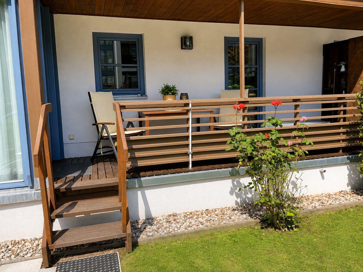 Terrasse mit Tisch, Stühlen und Blumenbeet. Holzgeländer und Treppe führen zur Terrasse.