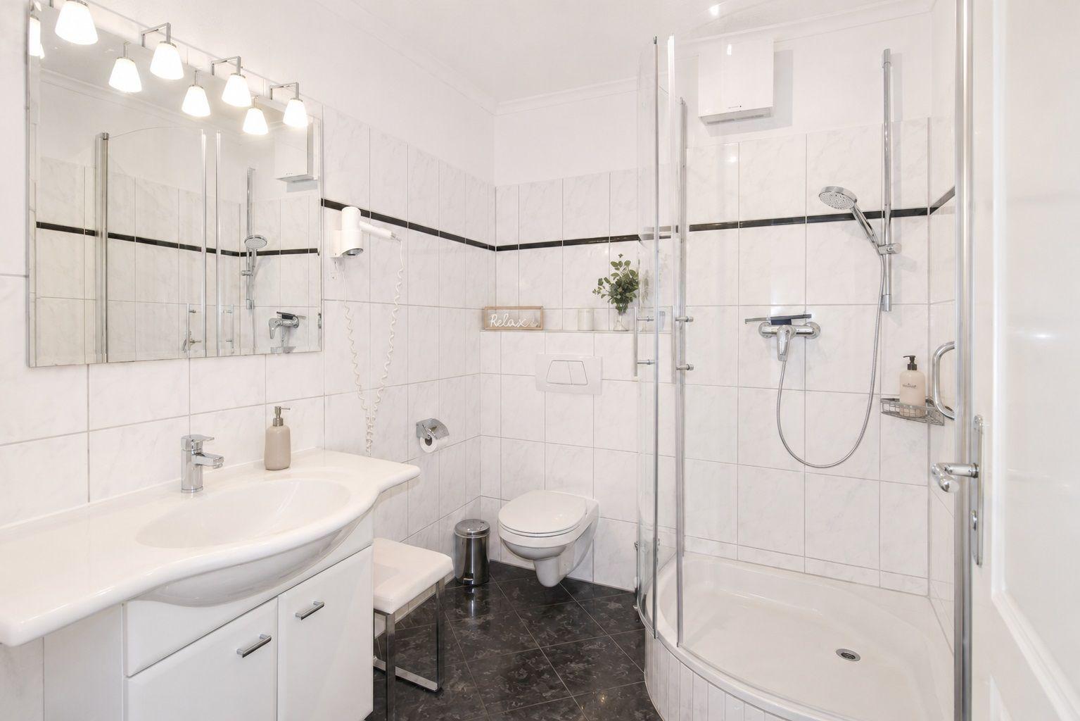 Badezimmer mit Dusche, Waschbecken und Toilette. Weiße Fliesen, dunkler Boden.