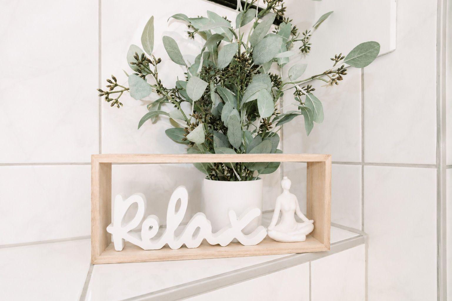 Holzrahmen mit Pflanze, Relax-Schrift und Meditationsfigur auf Fliesenwand.