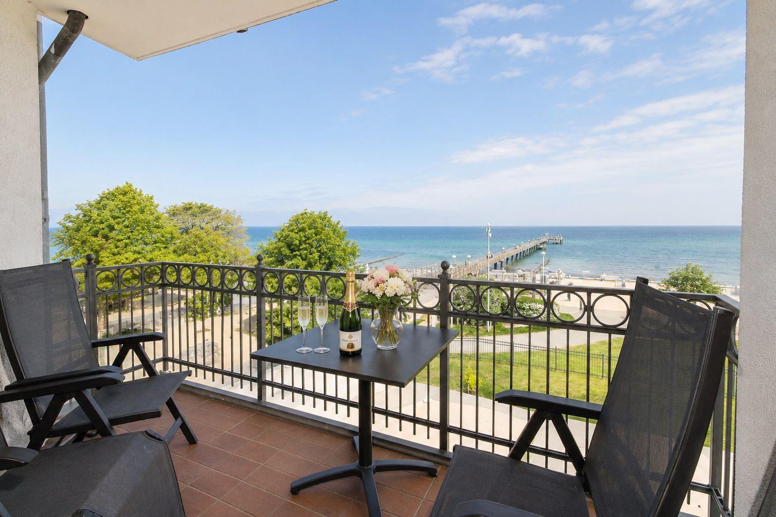 Terrasse mit Tisch, zwei Stühlen, Flasche Champagner und Blumen. Blick auf Meer und Pier.