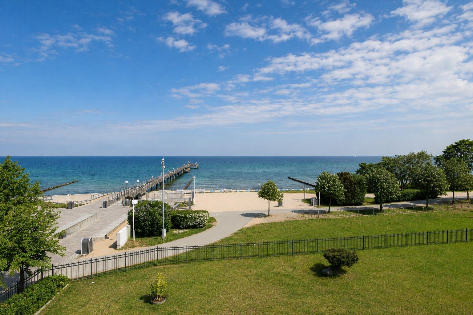 Strandpromenade mit Steg und grünen Flächen am Meer unter blauem Himmel.
