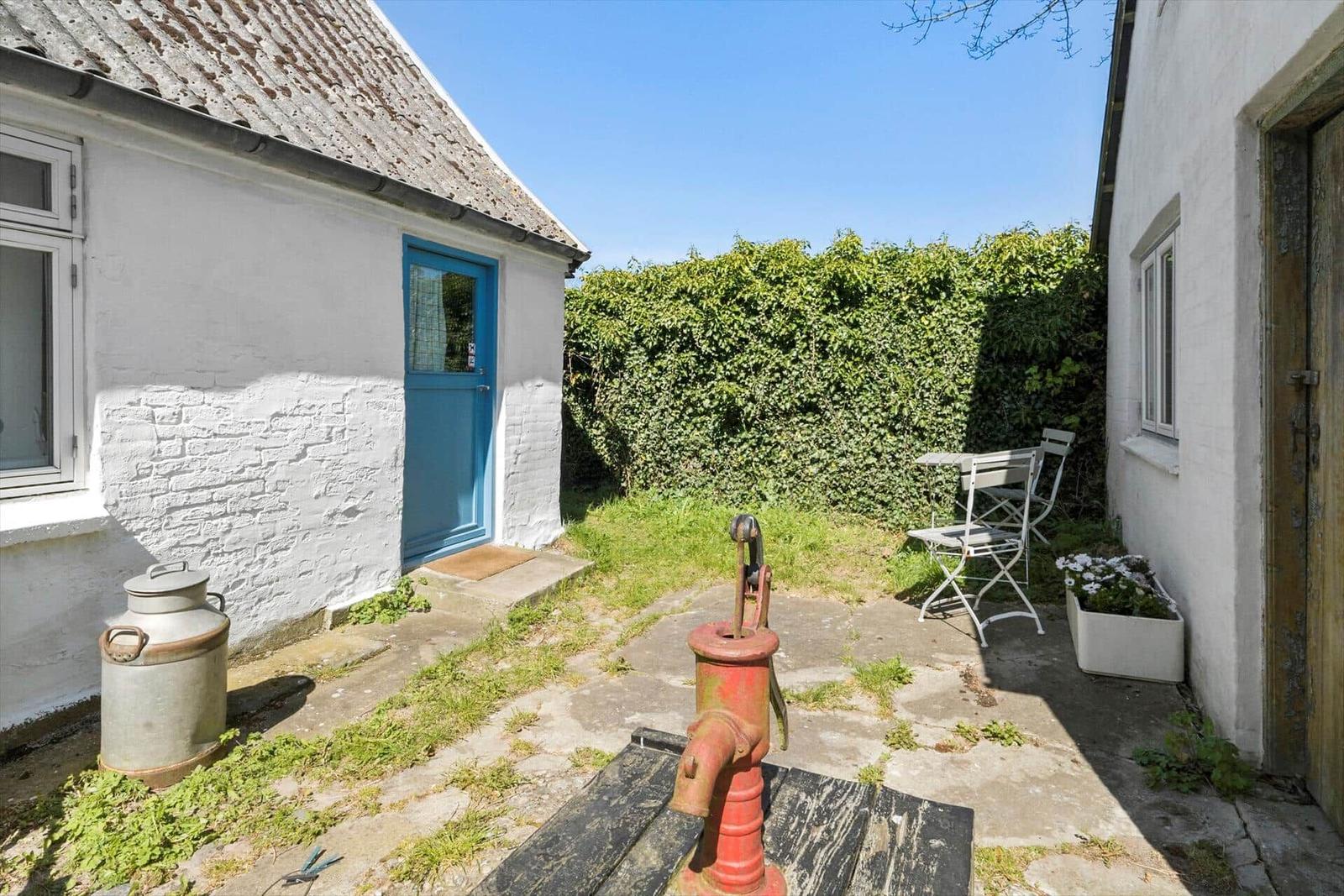 Garten mit Tisch und Stühlen, daneben Haus und Flagge. Bäume und Hecke im Hintergrund.