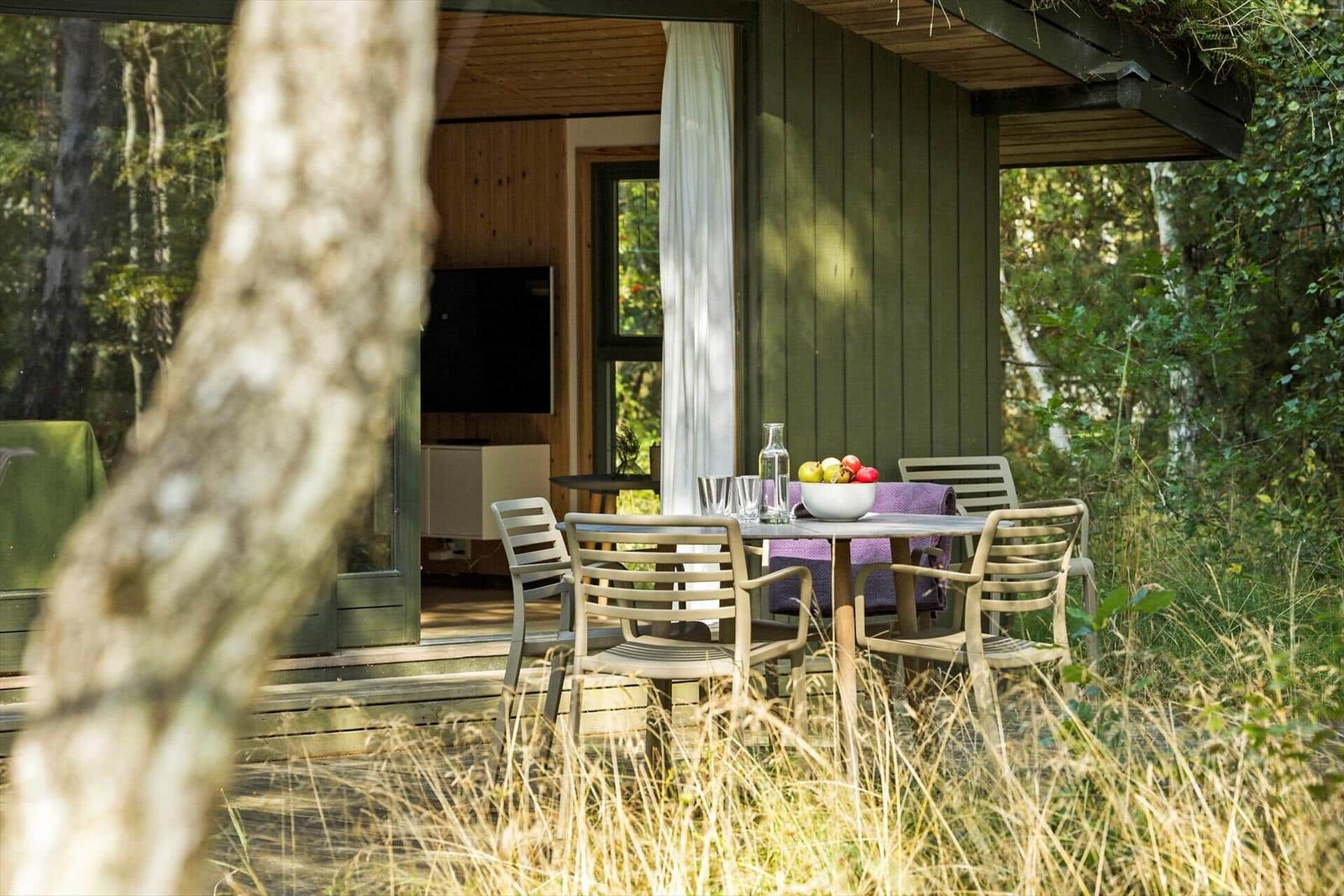 Holzhaus mit Grasdach und Terrasse im Wald. Grüne Stühle und Blumen auf der Veranda.