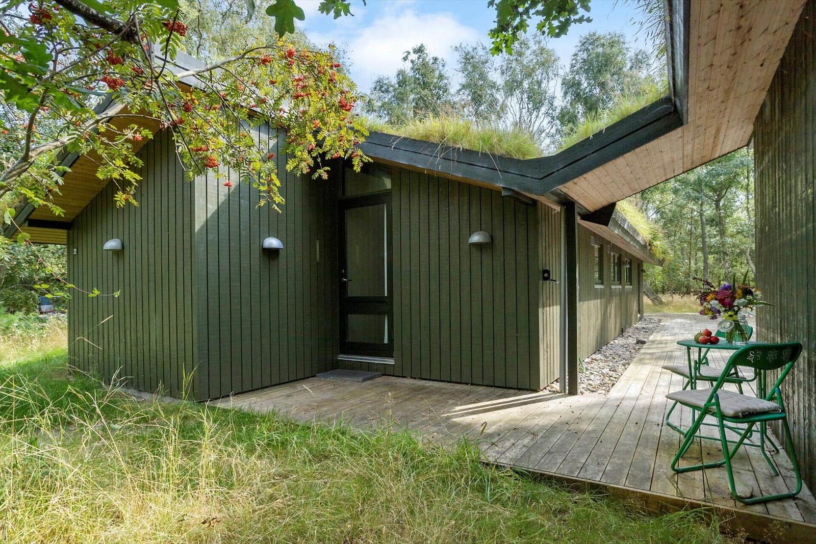 Holzterrasse mit Grillplatz und Sitzgelegenheit vor grünem Holzhaus mit Grasdach.