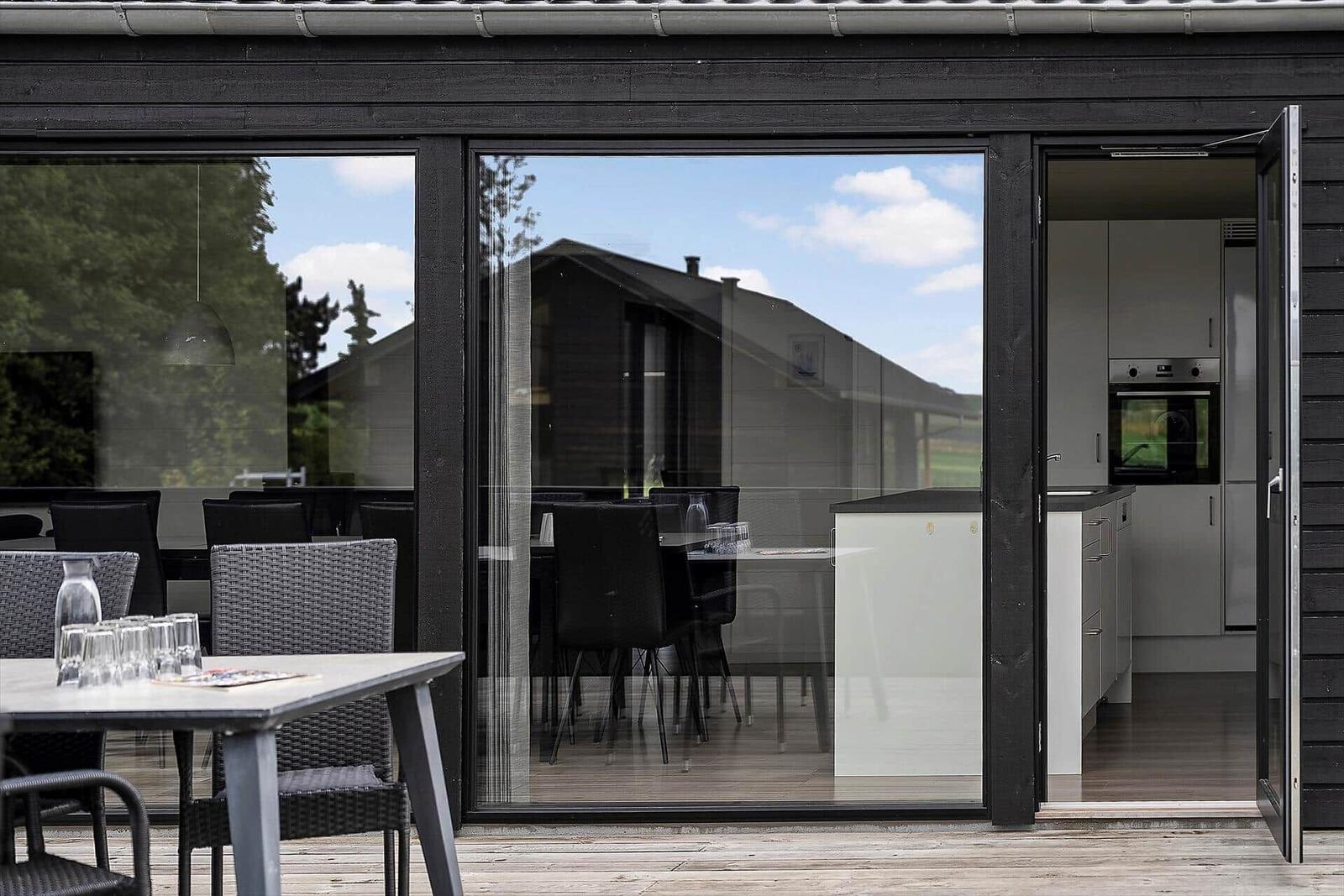 Modern villa with large wooden deck and seating. Glass wall connects indoor and outdoor areas.