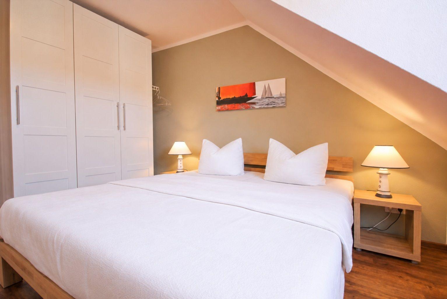 Schlafzimmer mit Dachschrägen, rotem Bettbezug, zwei Nachttischen und Wandbild.