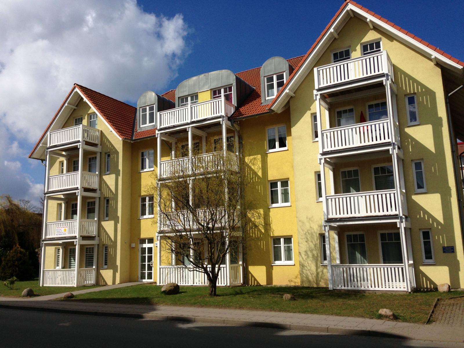Gelbes Mehrfamilienhaus mit weißen Balkonen und rotem Dach unter blauem Himmel.