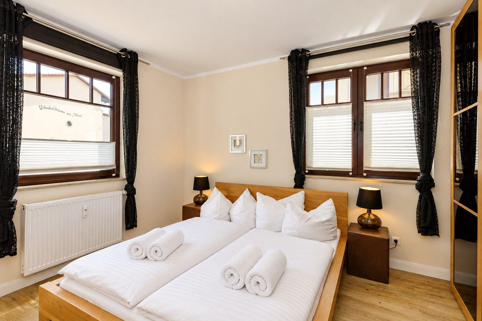 Schlafzimmer mit Doppelbett, zwei Fenstern und schwarzen Vorhängen.