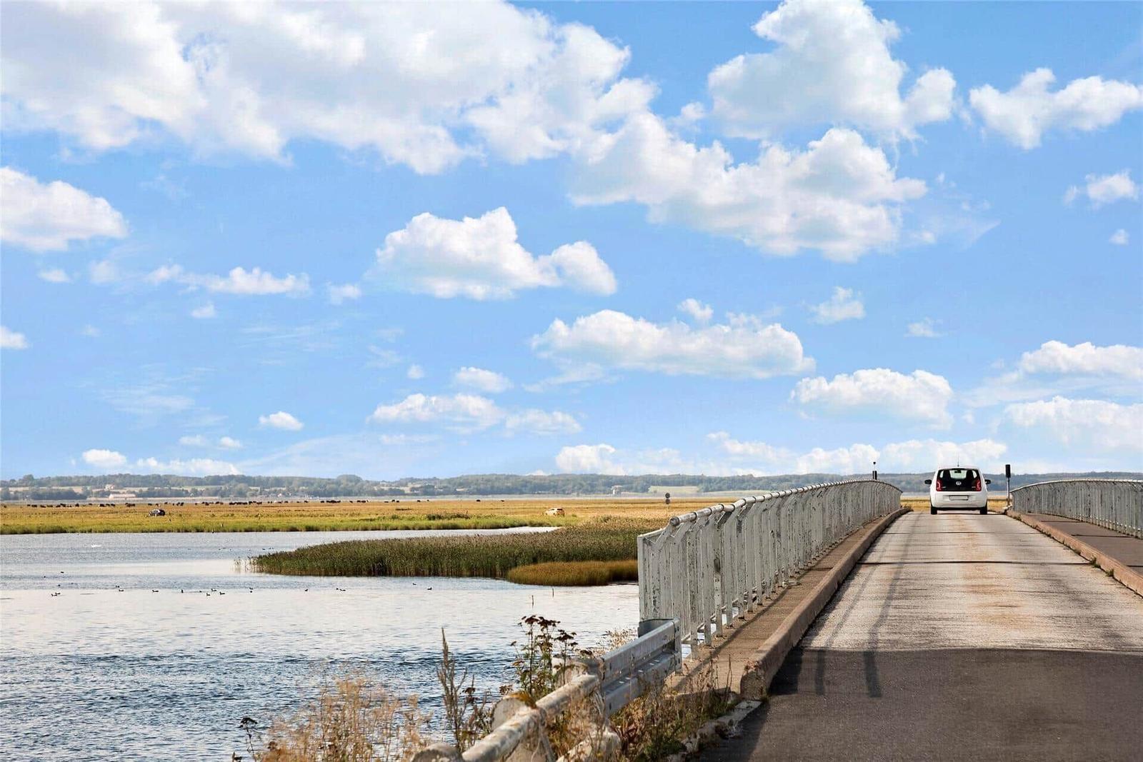 Ein weißes Auto fährt über eine Brücke über einen See mit Schilf und einem Himmel mit weißen Wolken.