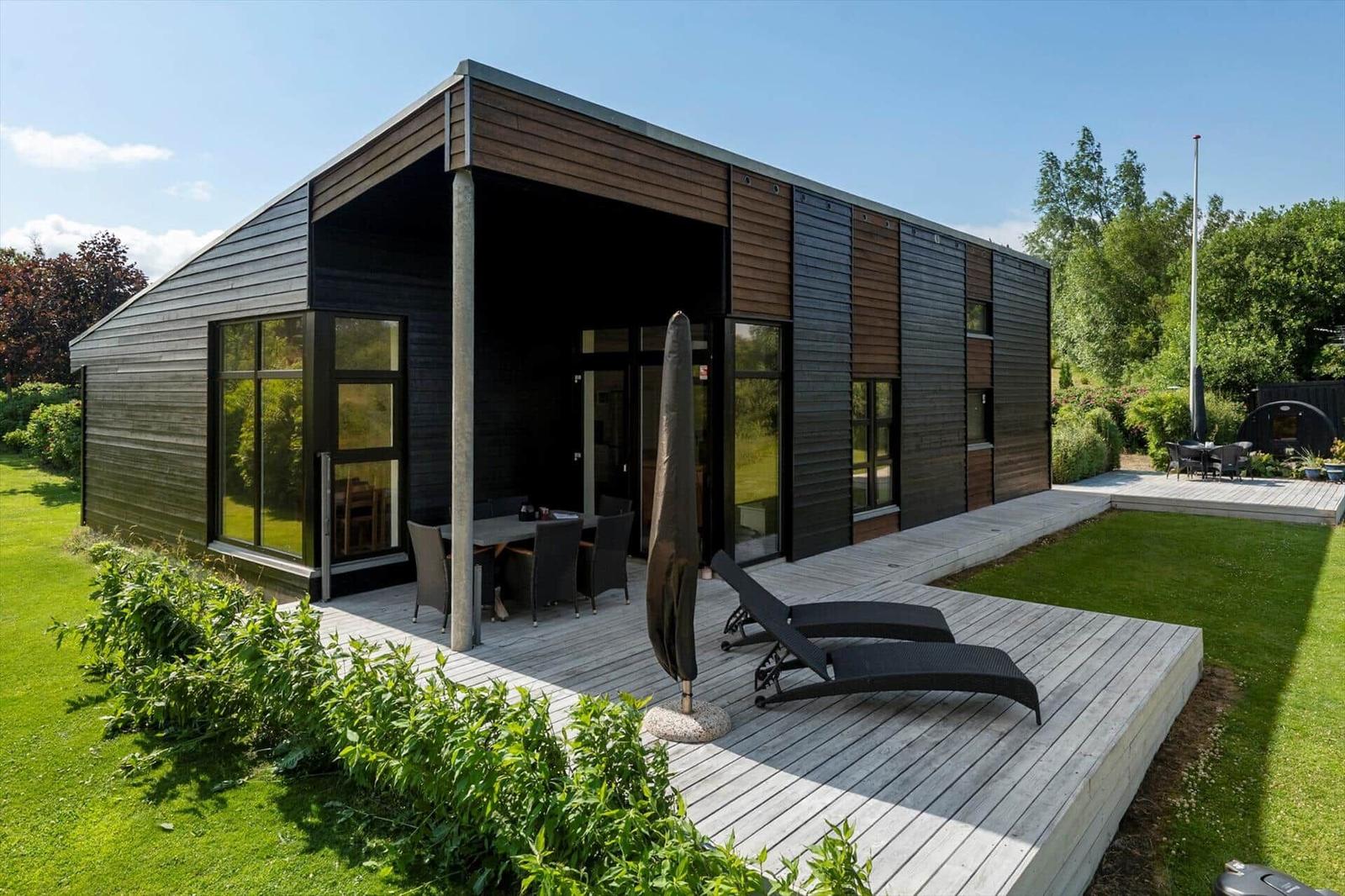Modern black cabin with terrace, surrounded by greenery and a lake.