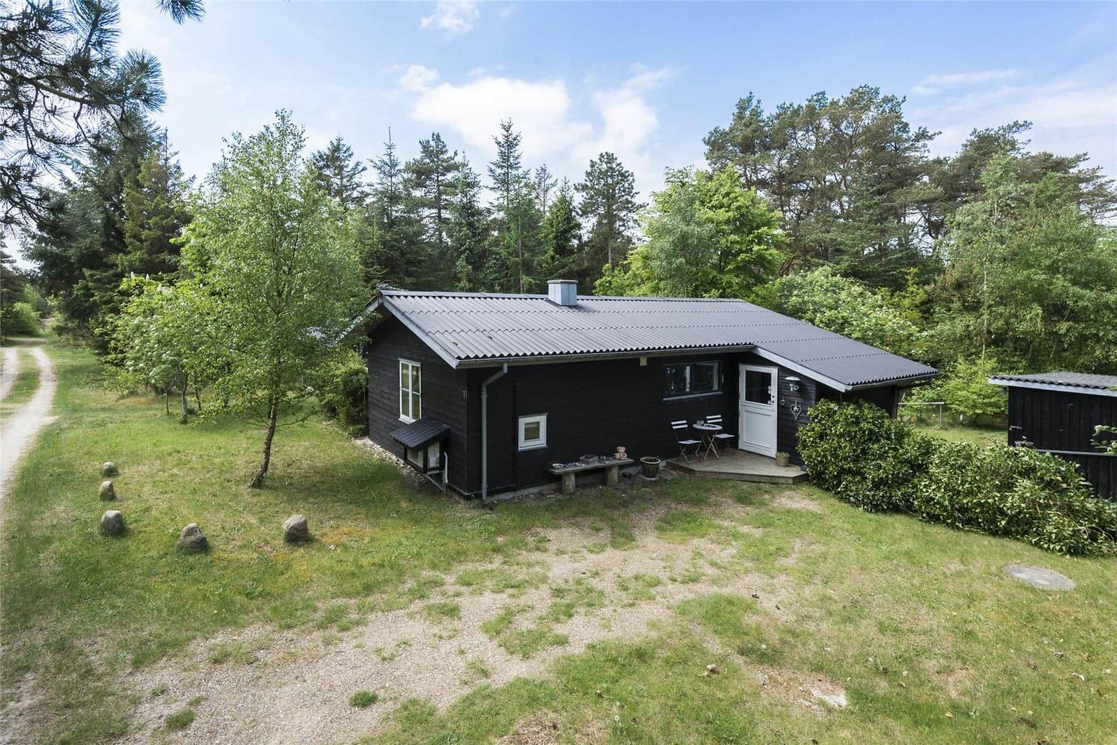 Holzterrasse mit Tisch und Stühlen vor einem schwarz gestrichenen Haus mit Garten und Bäumen.