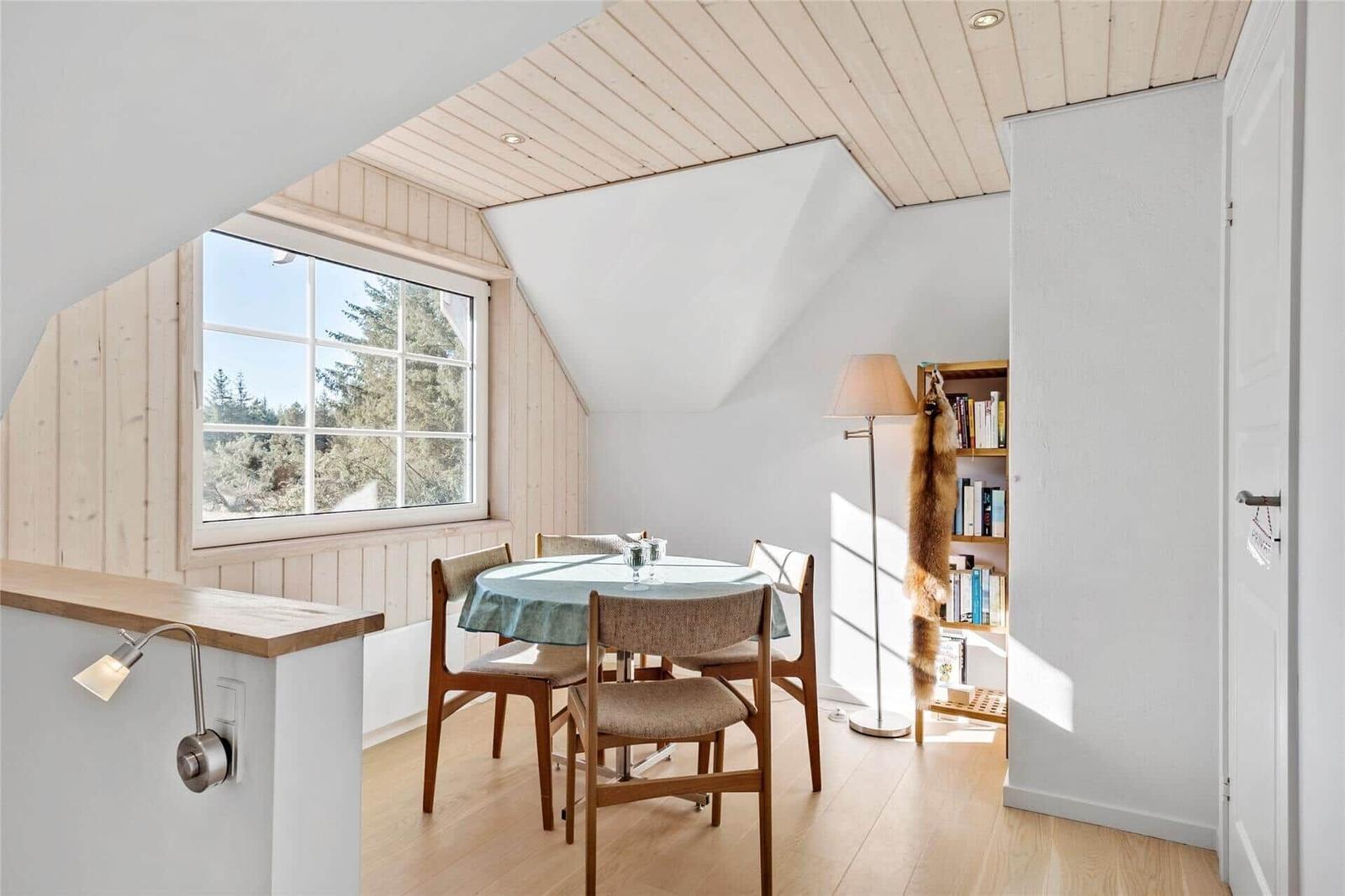 Esszimmer mit runden Tisch, vier Stühlen und großem Fenster. Holzdecke und Bücherregal sichtbar.