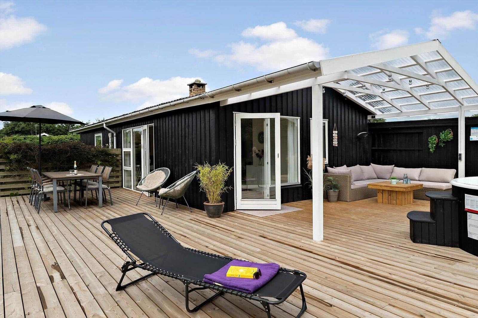 Backyard with hot tub, seating bench, and covered area. Black wooden wall and deck.