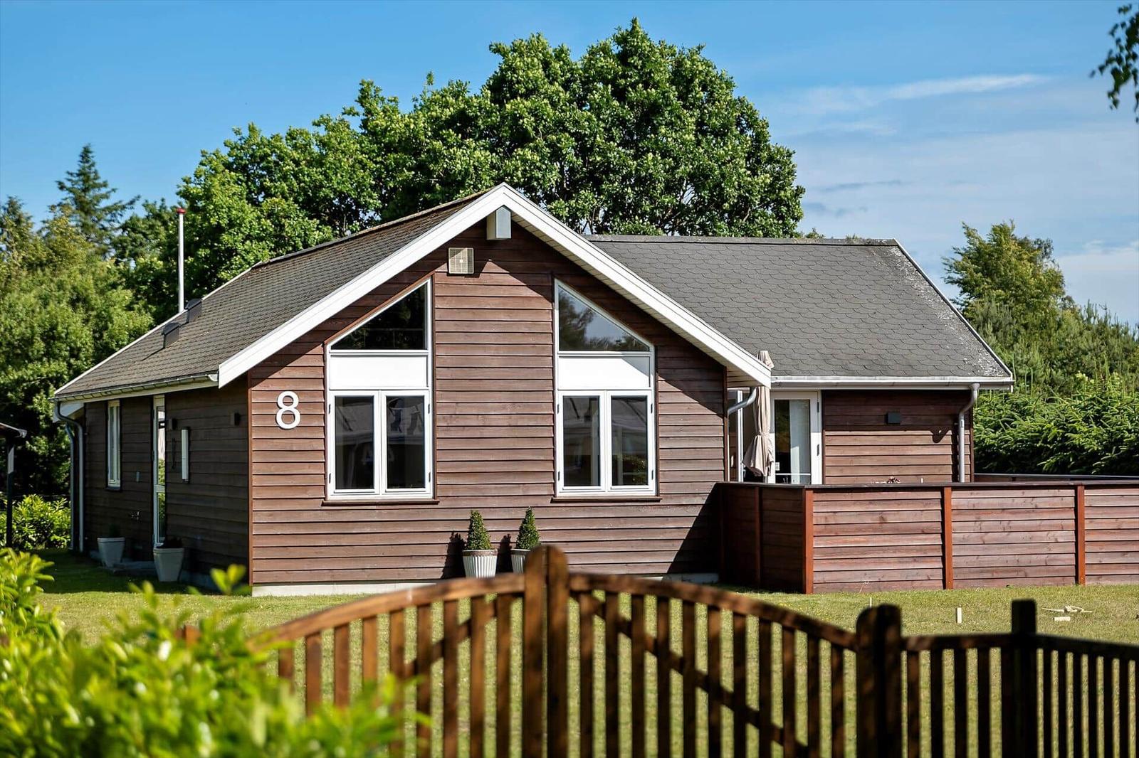 Haus mit Holzverkleidung, Terrasse und Garten. Große Fenster, Außentisch und Sitzgelegenheiten.