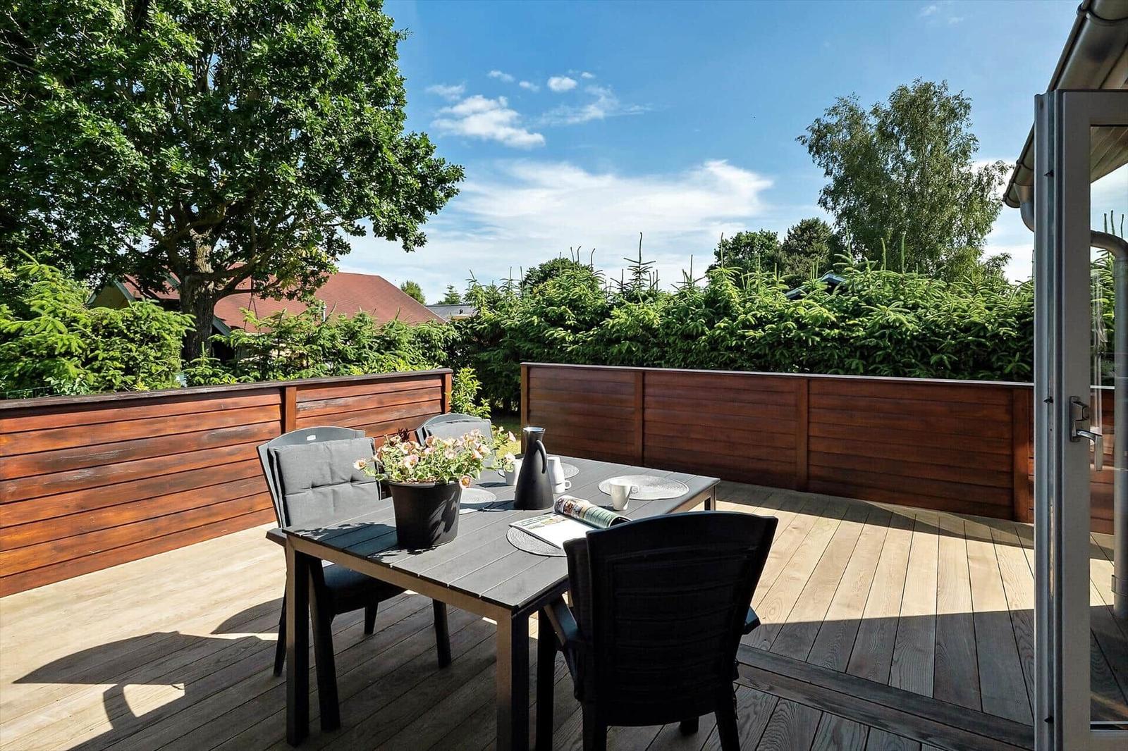 Terrasse mit Tisch, Stühlen, Grill und Pflanzen. Holzgeländer und Bäume im Hintergrund.