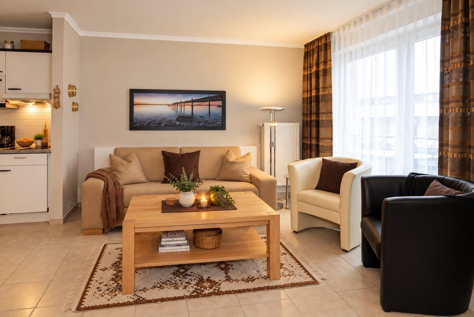 Wohnzimmer mit Sofa, Sessel und Couchtisch. An der Wand hängt ein Bild. Links ist die Küche.