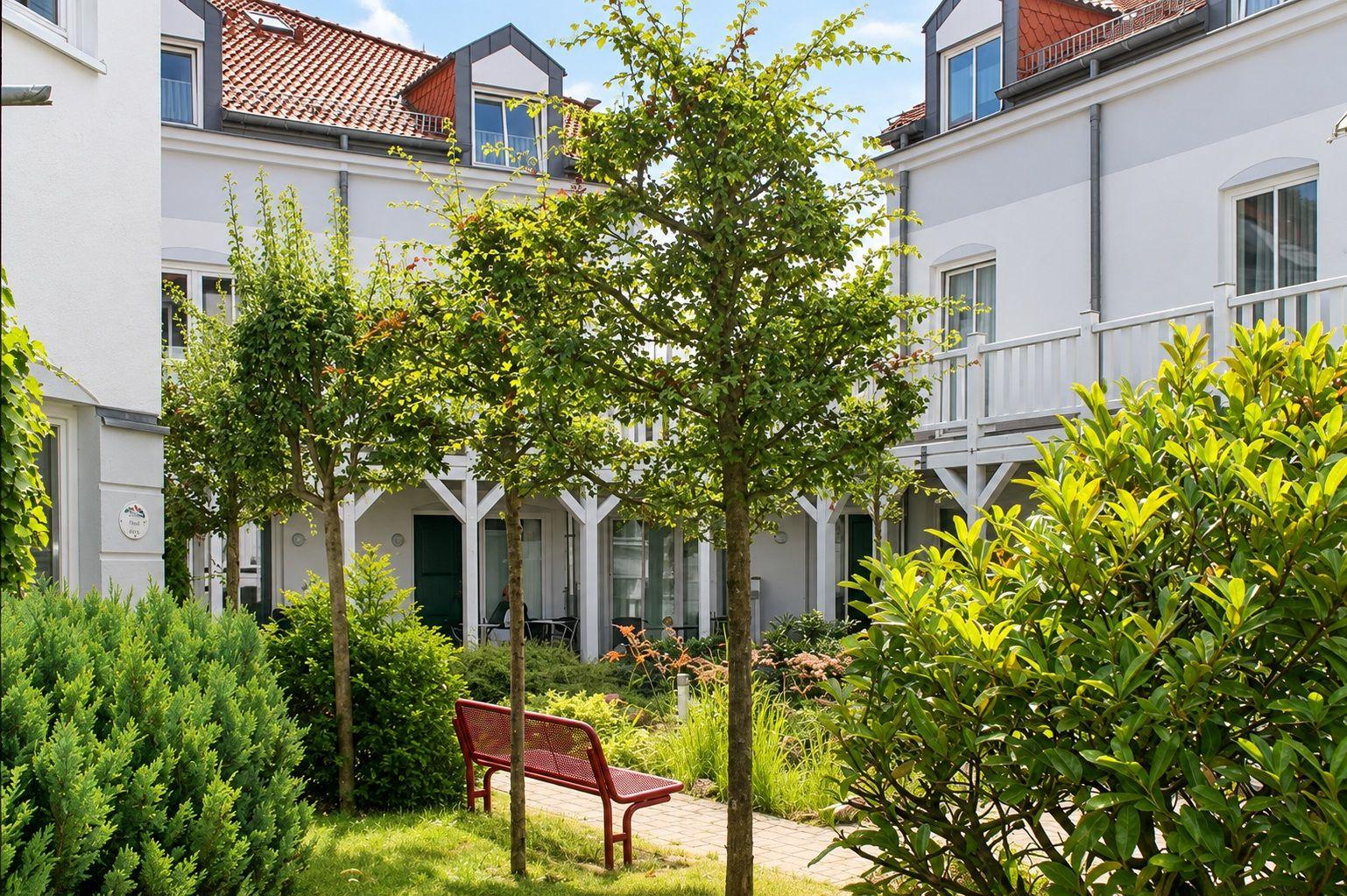 Hinterhof mit Bäumen, Beete und einer roten Bank zwischen weißen Häusern.