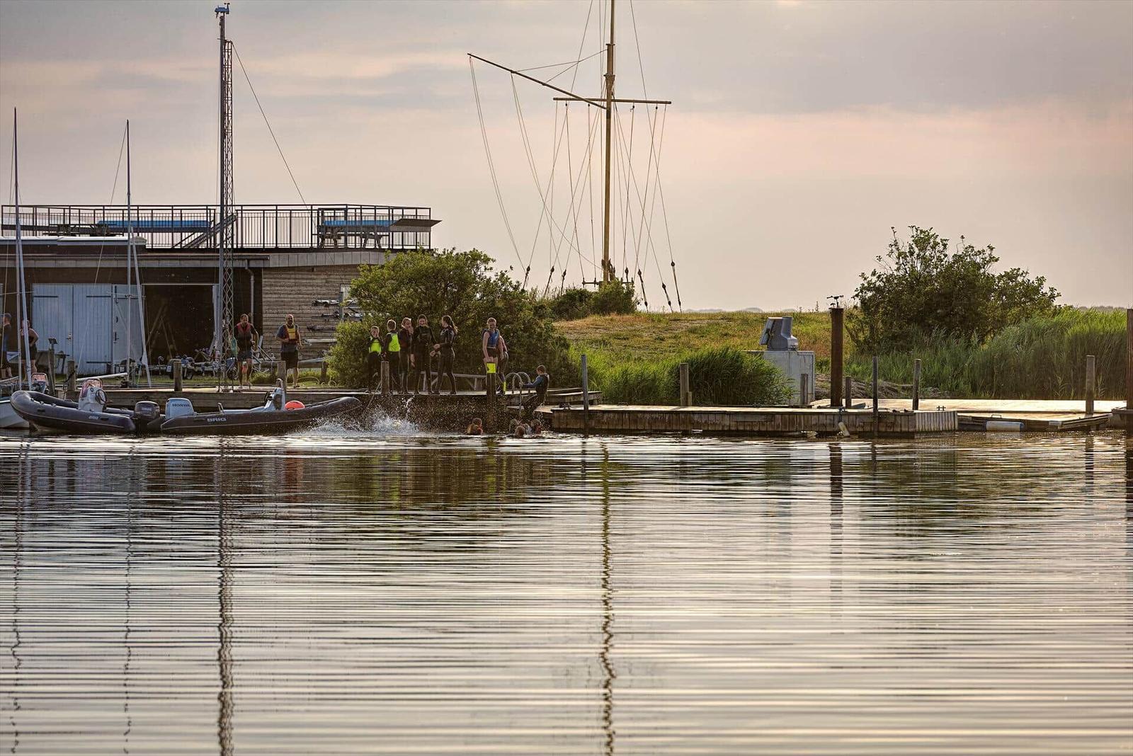Boot mit Motor auf Wasser, Menschen am Steg, Segelyacht im Hintergrund.