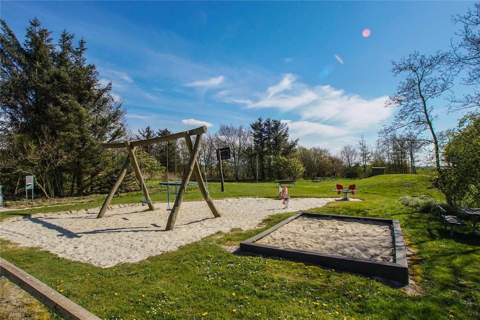 Spielplatz mit Sandkasten, Schaukel und Rutsche im grünen Freigelände.