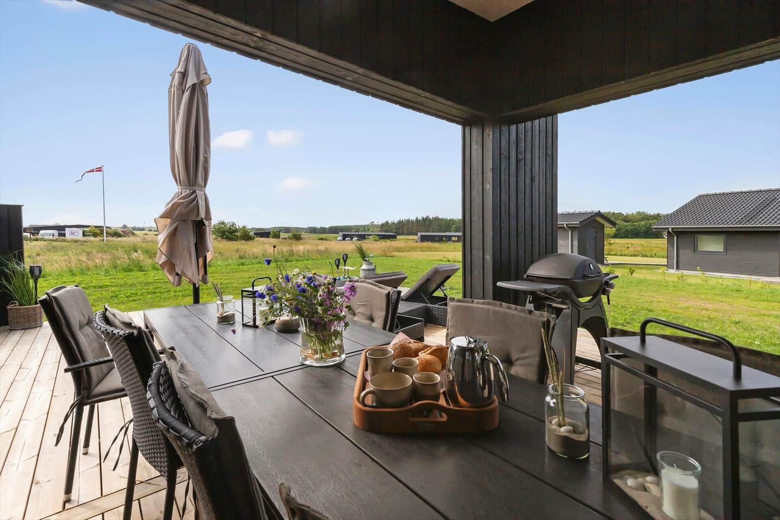 Holzterrasse mit schwarzer Umzäunung und Gartenmöbeln. Im Hintergrund sind Häuser und Wiesen zu sehen.