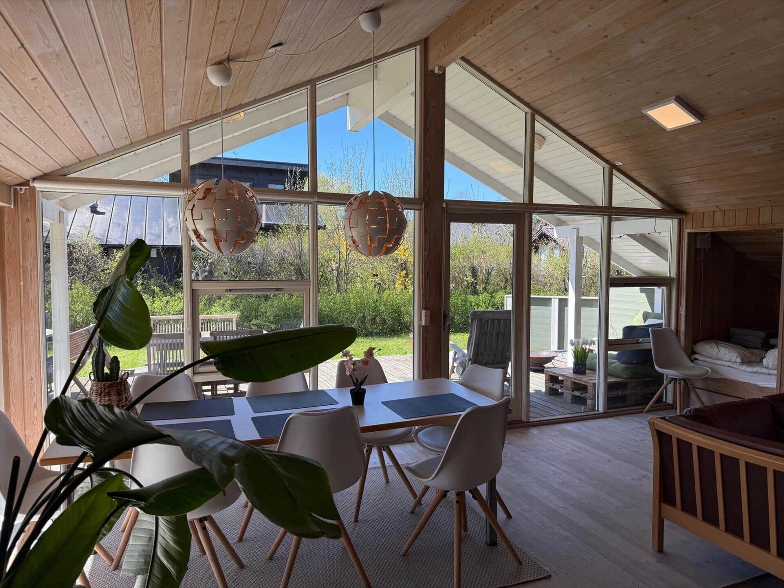 Dining area with table, chairs, and large windows. Wooden walls and ceiling. Plants and pendant lights.