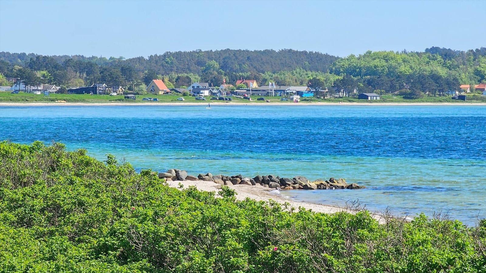 Beach with clear water and green vegetation in the foreground. Houses and trees are visible in the background.