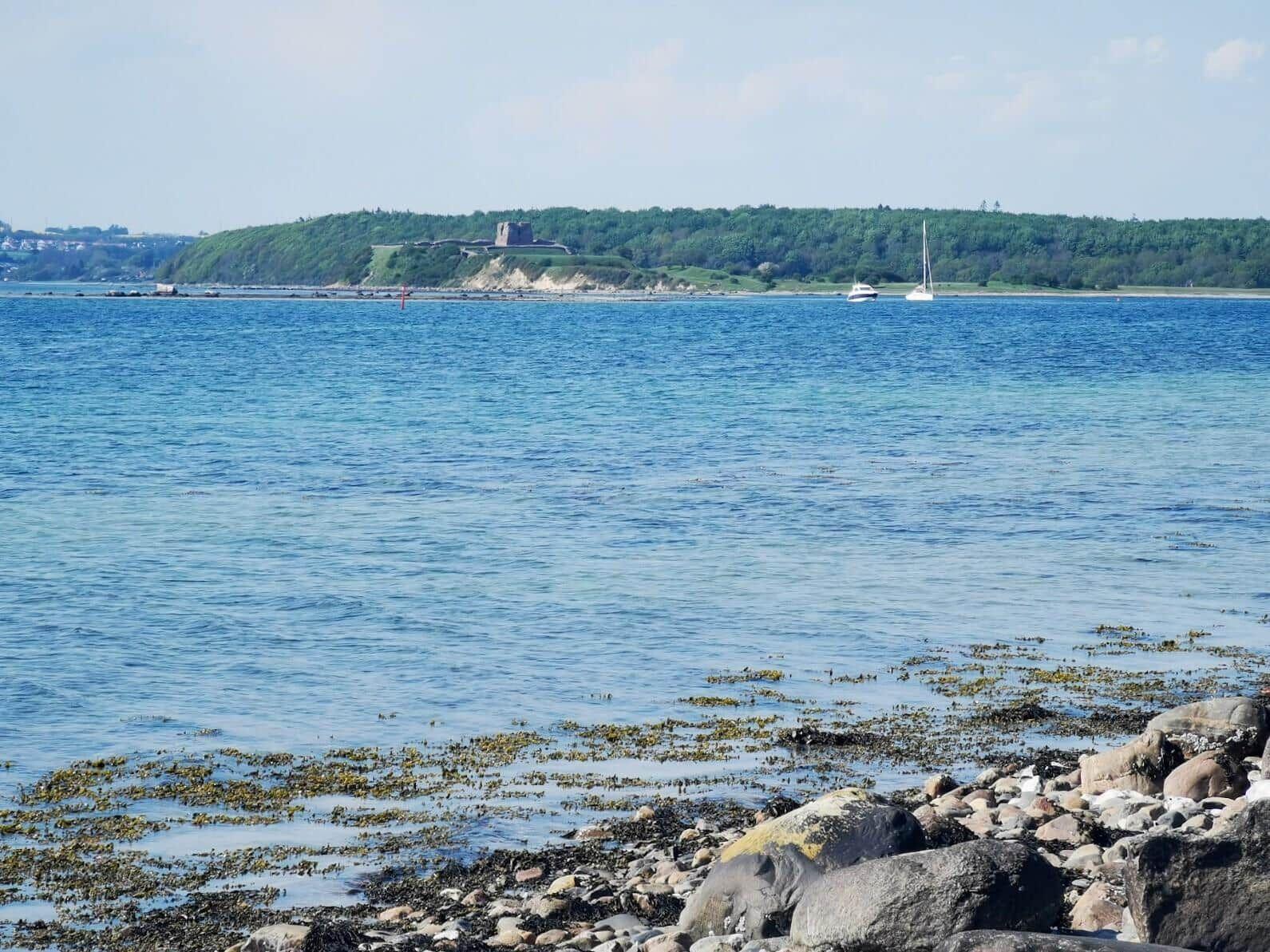 Coastal view with rocks, seaweed, and boats before a green island with a castle.