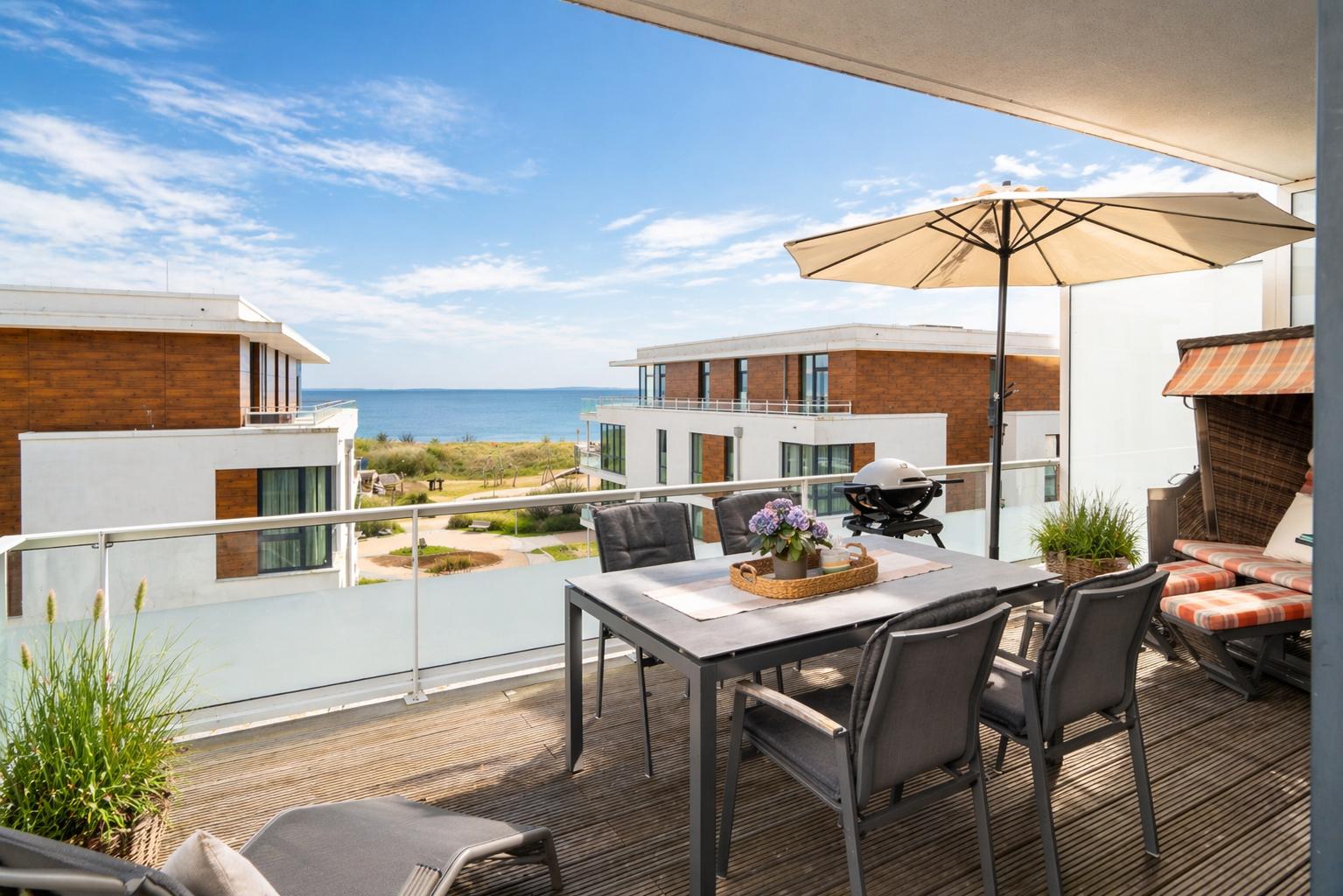 Terrace with table, chairs, umbrella, and grill. View of modern buildings and beach.