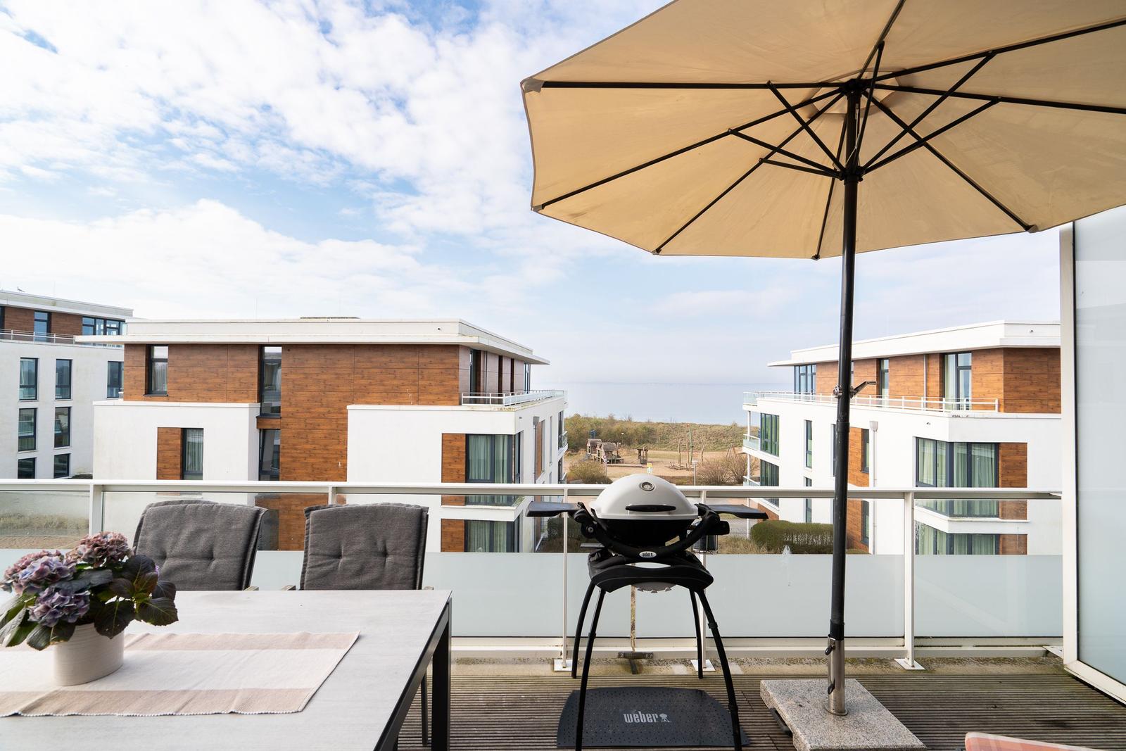 Terrace with table, chairs, umbrella, and lounge chair. View of residential buildings and sea.