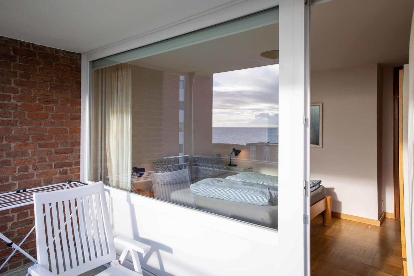 Schlafzimmer mit Blick aufs Meer durch Glastür und Balkon mit weißen Stühlen.