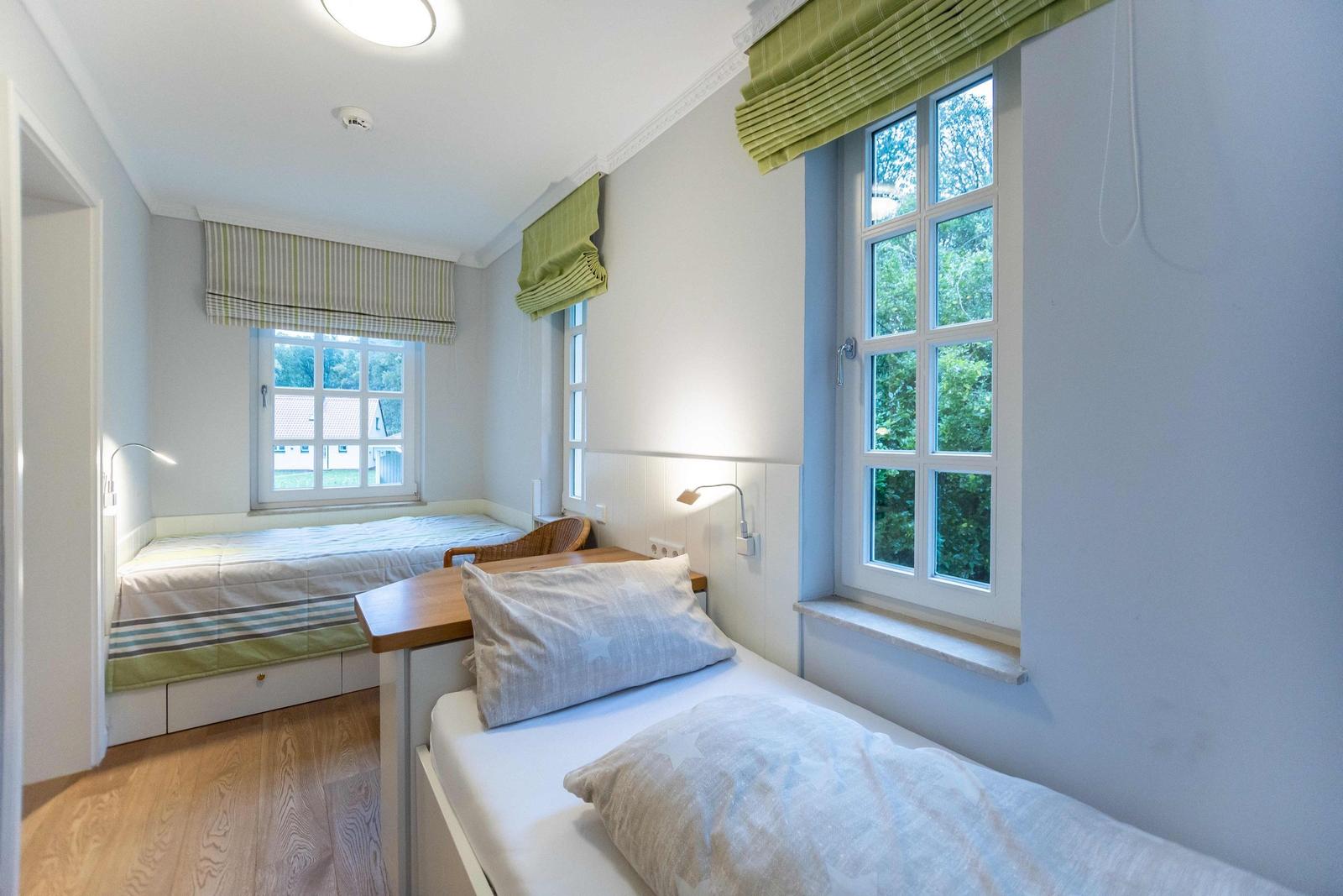 Ein Schlafzimmer mit Bett, Tisch und zwei Fenstern mit grünen Rollläden.