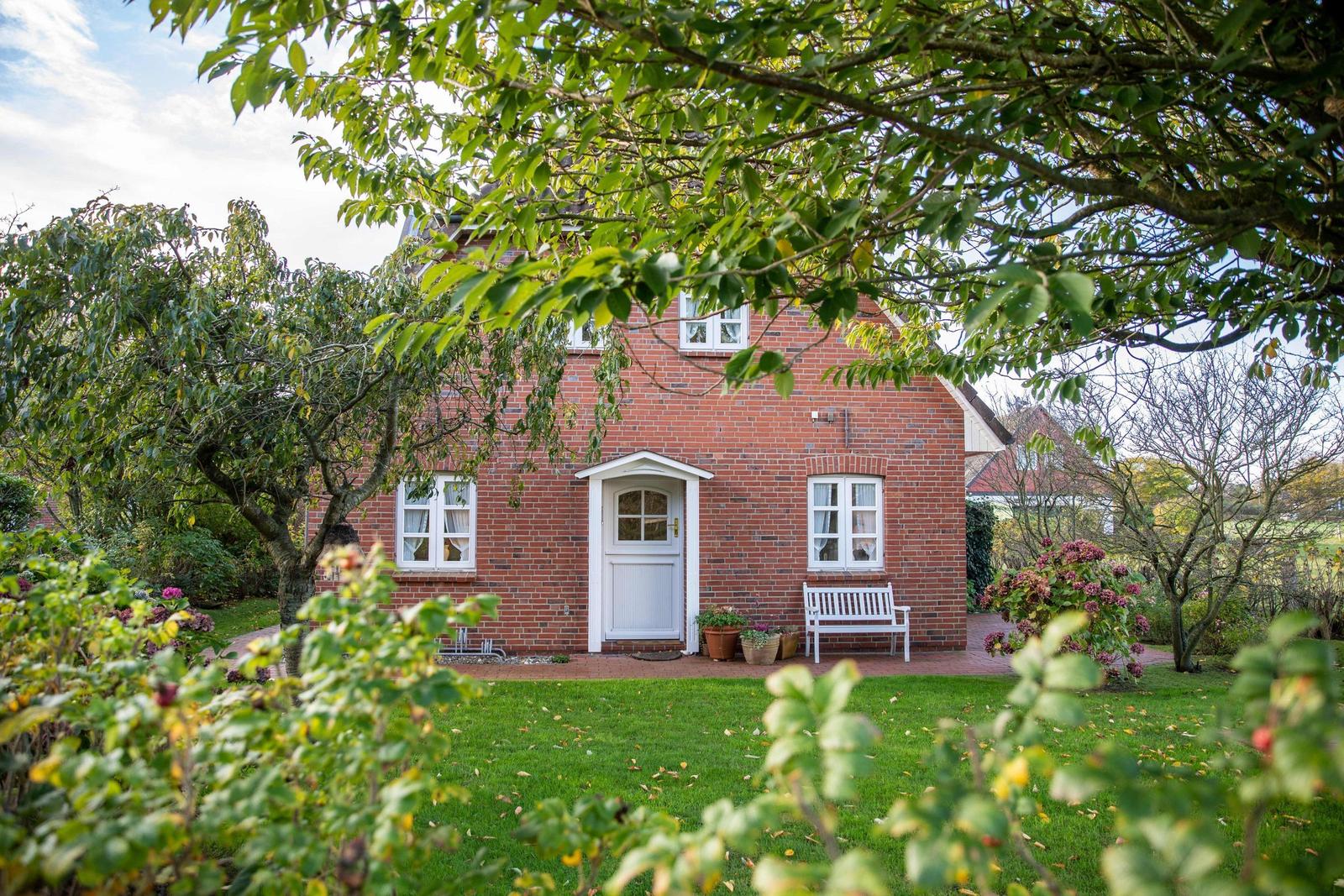Haus mit Garten, Bäumen und Blumen. Rasenfläche und Steinmauer sichtbar.