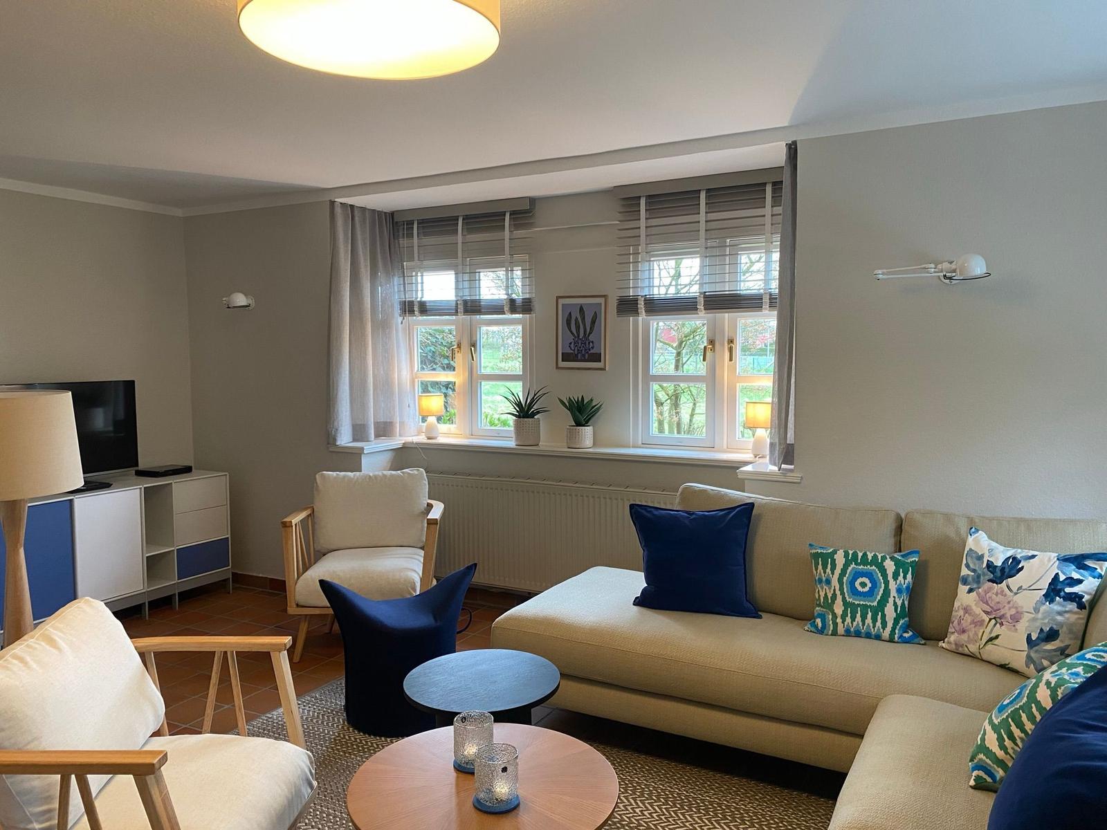 Wohnzimmer mit Sofa, Sessel und Fenstern. Auf dem Fensterbrett stehen Pflanzen.