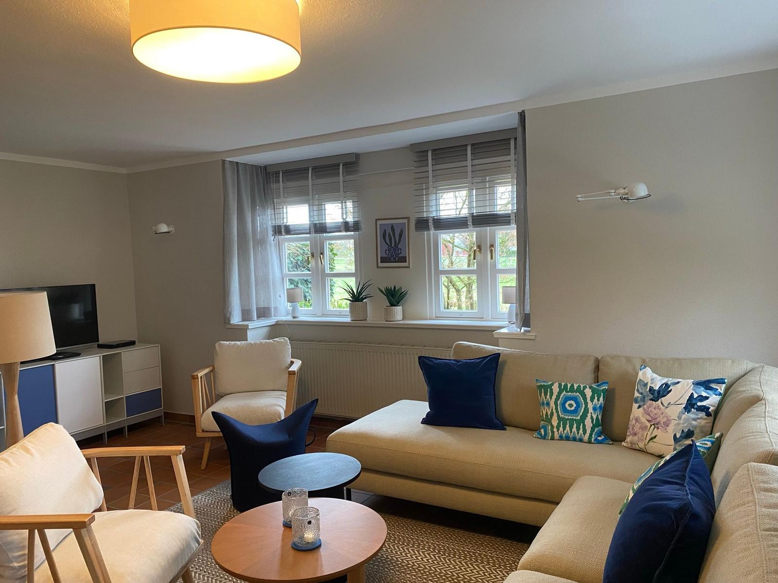 Wohnzimmer mit Sofa, Sesseln und Fenstern. Auf dem Fensterbrett stehen Pflanzen.