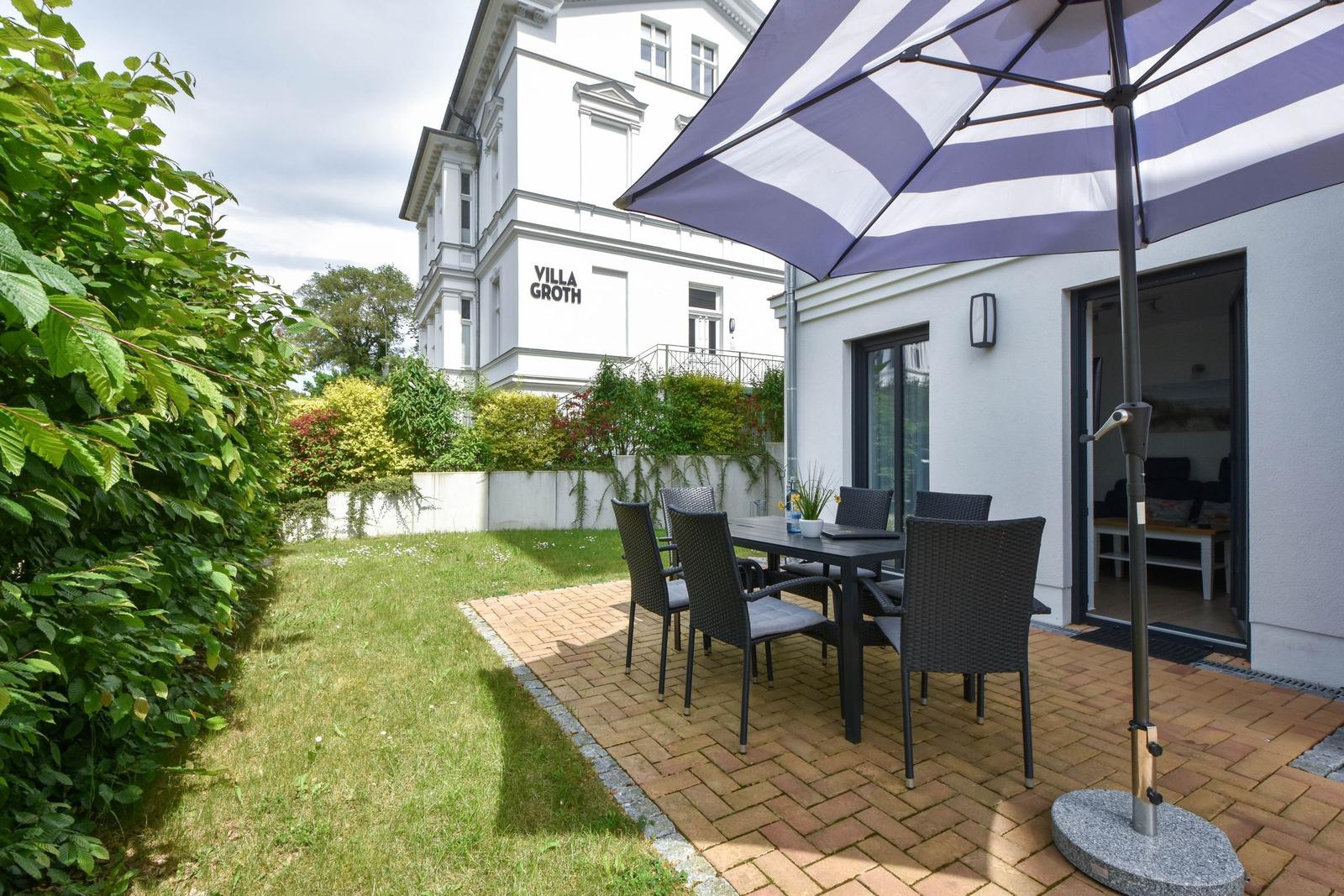 Terrasse mit Tisch, Stühlen und Sonnenschirm. Grünhecke und gepflasterte Fläche.