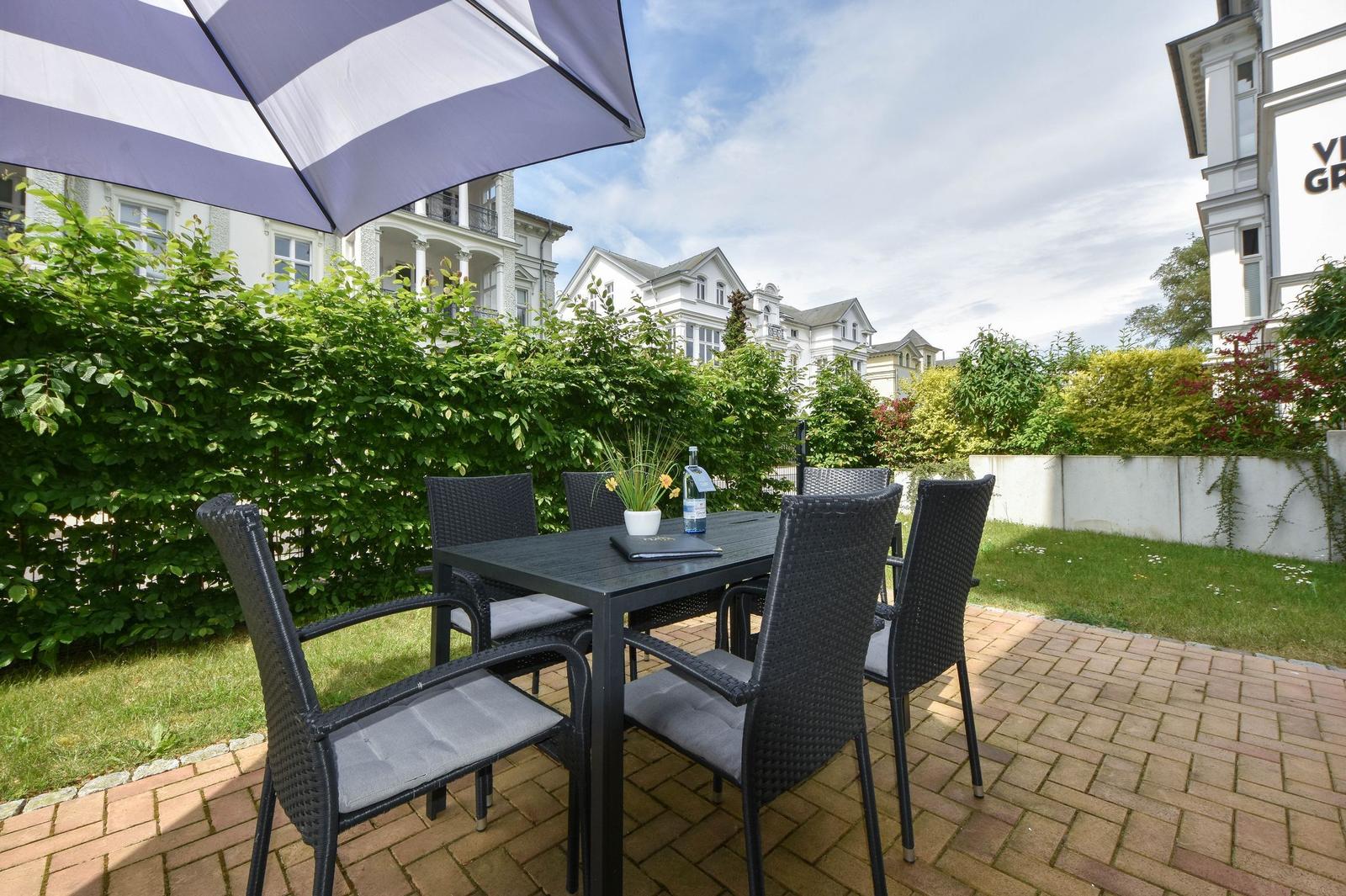 Terrasse mit Tisch, Stühlen und Sonnenschirm. Hinterhof mit Grünflächen und Villa Groth.