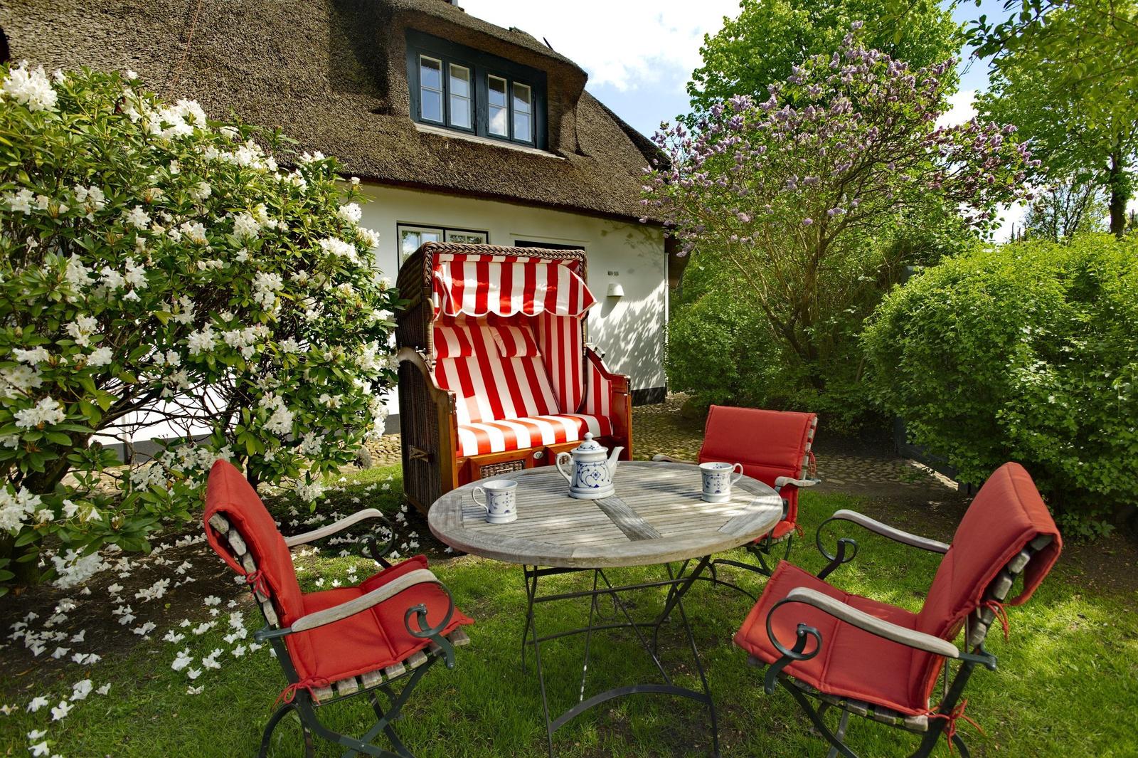 Garden with wicker chair, table and chairs in front of a thatched roof house.