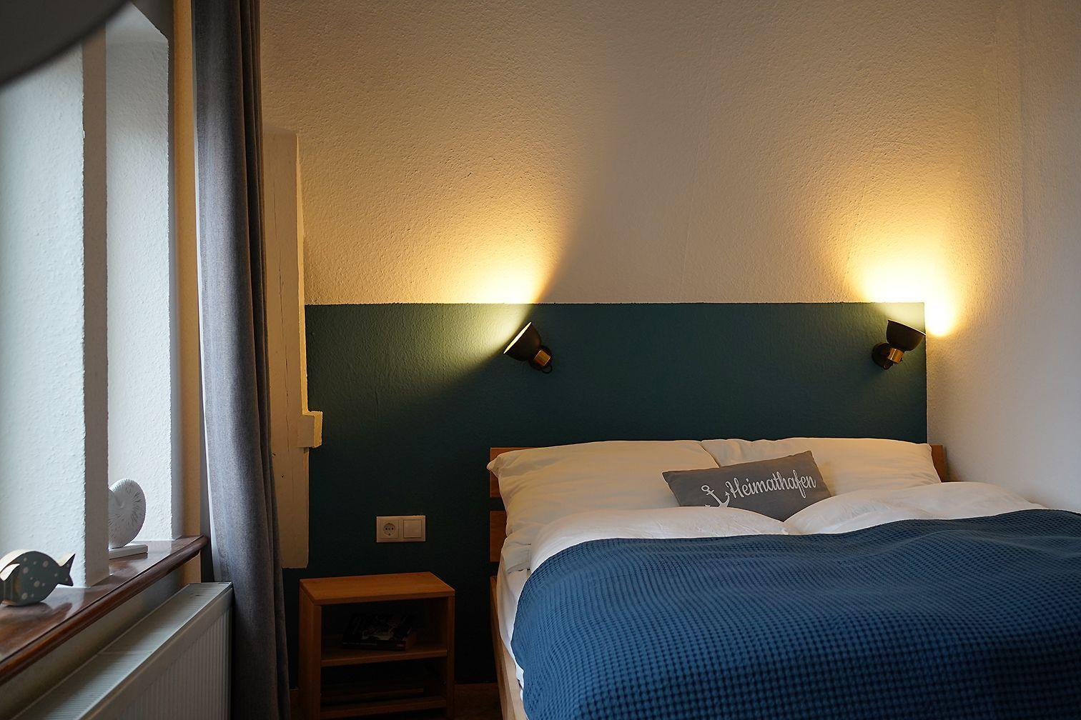 Ein Schlafzimmer mit blauer Sitzgelegenheit, Holzkommode und Stehlampe neben einem Fenster mit grauen Vorhängen.