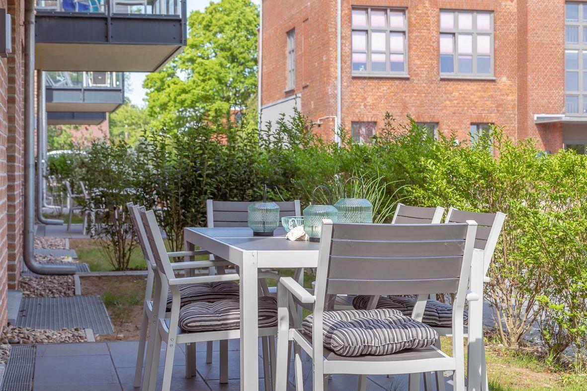 Terrasse mit Tisch, Stühlen und grünen Pflanzen. Glasfront und Backsteinmauer sichtbar.