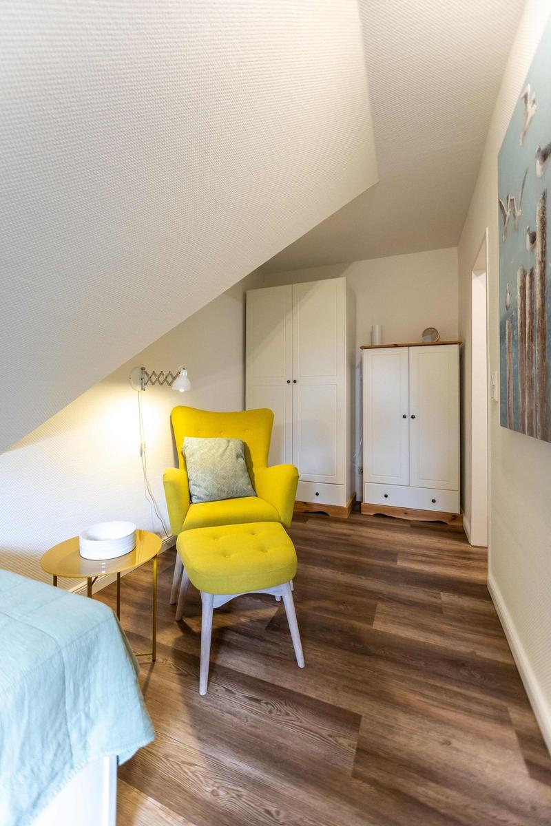 Schlafzimmer mit gelbem Sessel, Bett und weißen Schränken unter schrägem Dach.