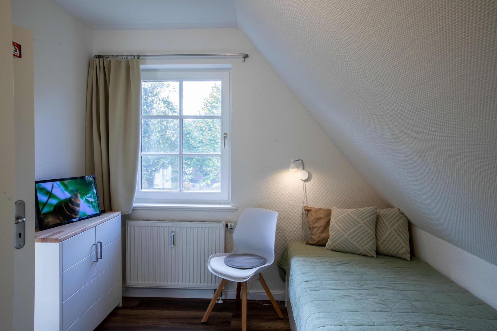Schlafzimmer mit Bett, Sessel und Fenster. Holzboden und Wandlampen.
