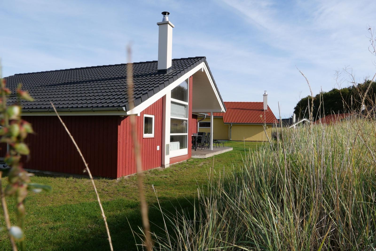 Roter Holzhaus mit Terrasse und Liegestühlen im Gras. Hintergrund: weitere Häuser und Himmel.