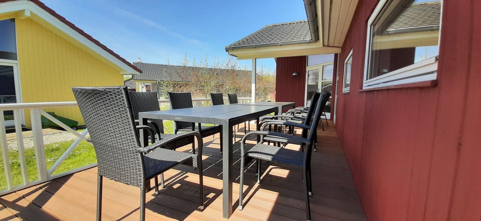 Rotes Ferienhaus mit Terrasse, Tisch und Stühlen. Grünfläche und klarer Himmel.